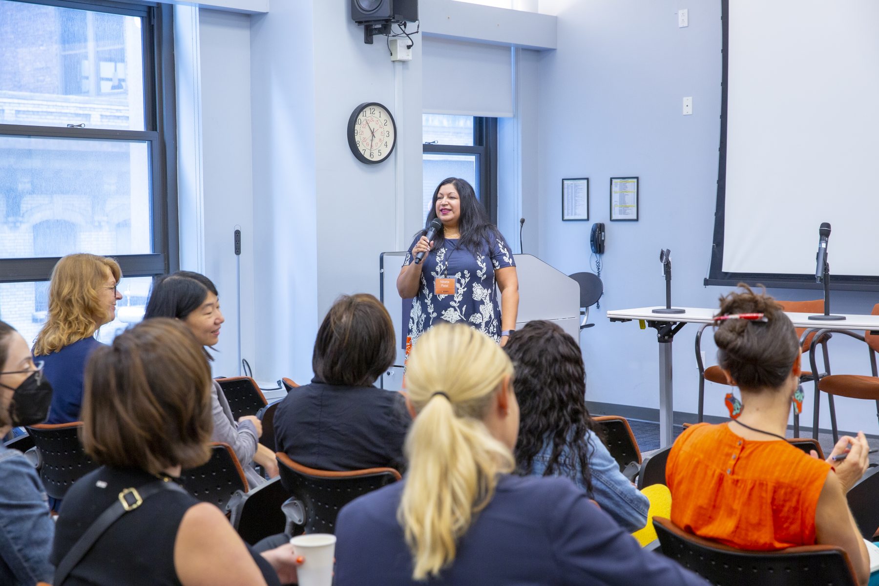 A woman in a navy blue floral dress stands at the front of a room, speaking into a microphone and smiling. She is addressing a seated audience. Behind her, there is a large screen and a table with microphones set up for a panel discussion.